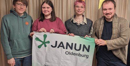 Vier Menschen mit janun-Banner lächeln in die Kamera. Text: Herzliche Einladung. Offenes Treffen des Vorstands am 22. Juli 26 von 16 Uhr bis 18 Uhr im janun-Büro (Bahnhofstr. 21). Oben rechts das Logo von janun Oldenburg.